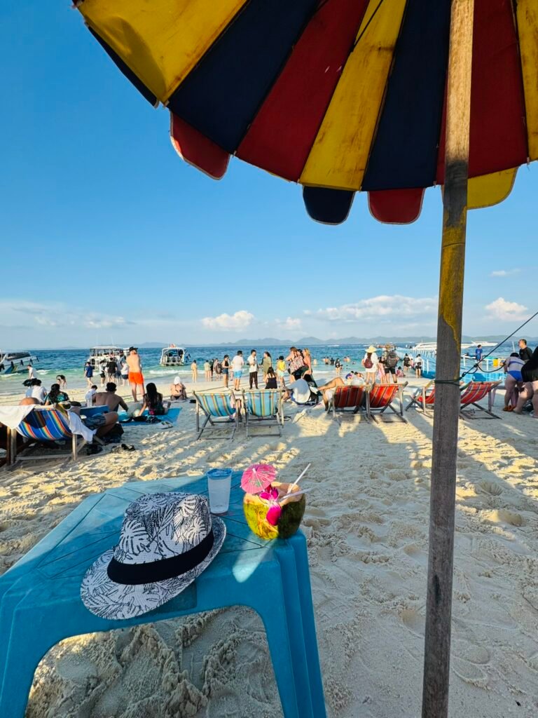 Beach scene at Khai Island with a hat and fresh coconut drink on a blue table under a colorful striped umbrella, beachgoers and boats visible in the background.
