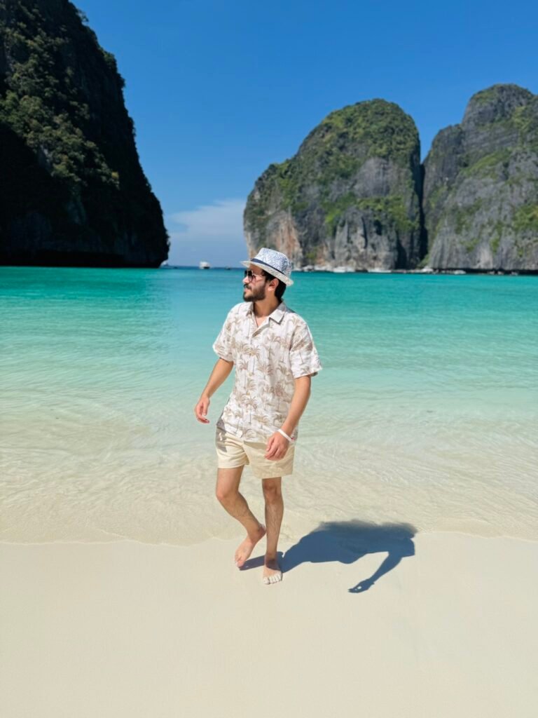 Man walking along the white sand shoreline at Maya Bay, Phi Phi Leh, with crystal-clear shallow water and dramatic limestone cliffs rising behind him.