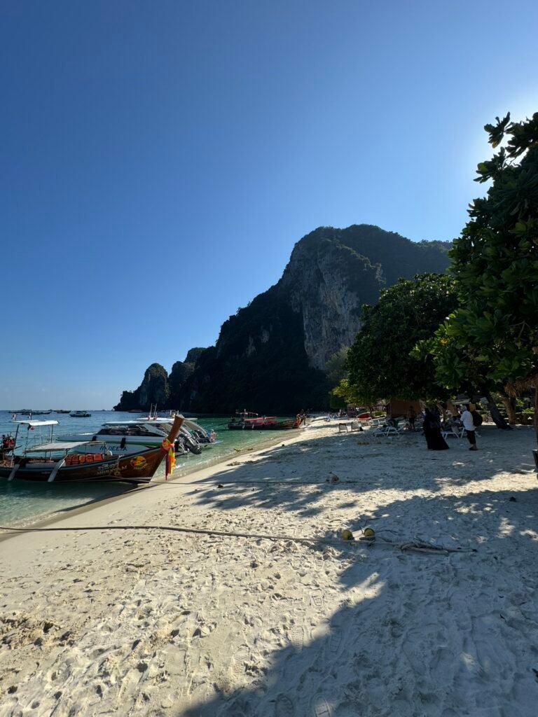 White sand beach on Phi Phi Don with longtail boats and speedboats moored along the shore, a limestone cliff in the background, and palm trees providing shade.