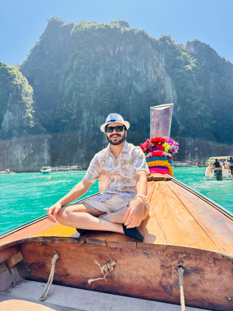 Man sitting cross-legged on the bow of a decorated Thai longtail boat with limestone cliffs and emerald water in the background at Phi Phi Islands.